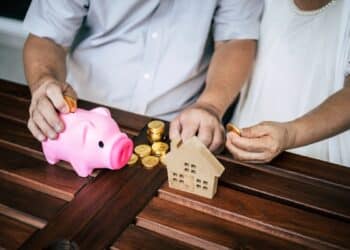 Casal economizando dinheiro com moedas, cofrinho rosa e miniatura de casa sobre a mesa.