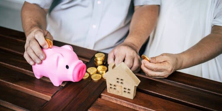 Casal economizando dinheiro com moedas, cofrinho rosa e miniatura de casa sobre a mesa.