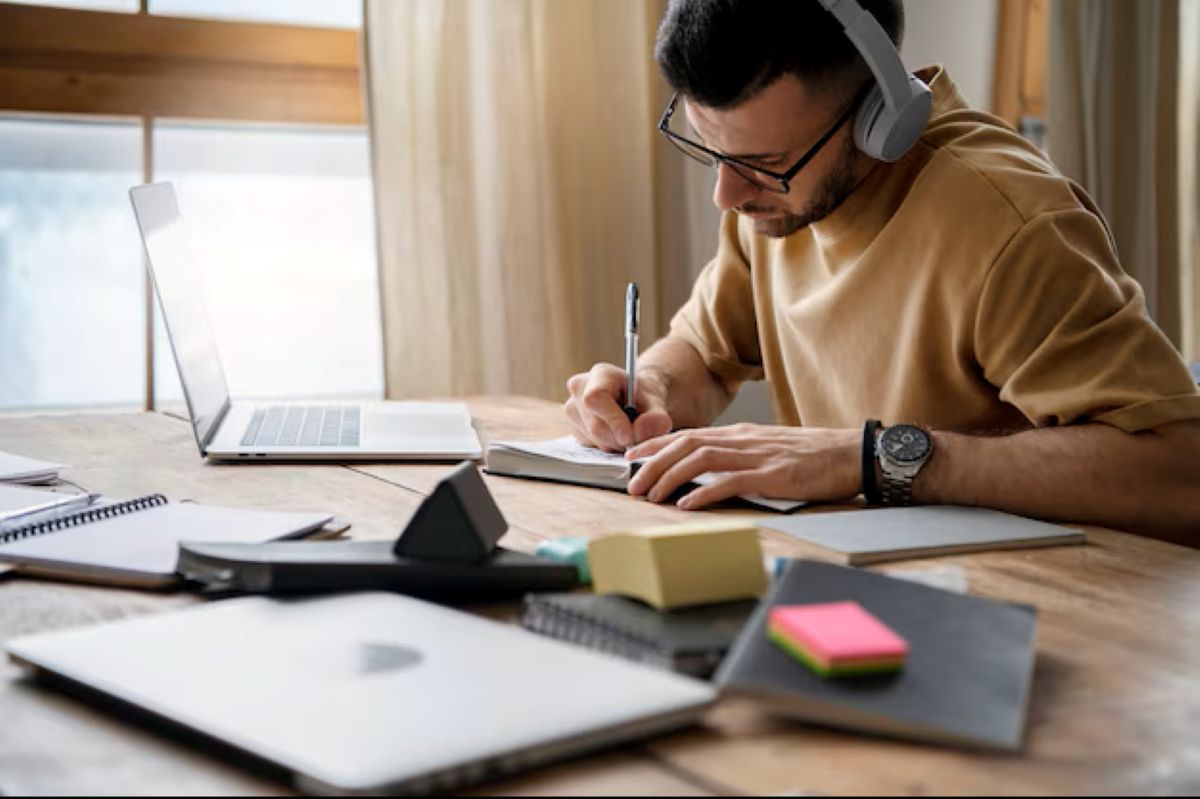 Homem estudando em casa para concurso do Banco do Nordeste com laptop e materiais de estudo.