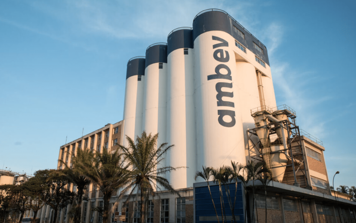 Fábrica da Ambev com silos brancos e logotipo da empresa em destaque sob céu azul.