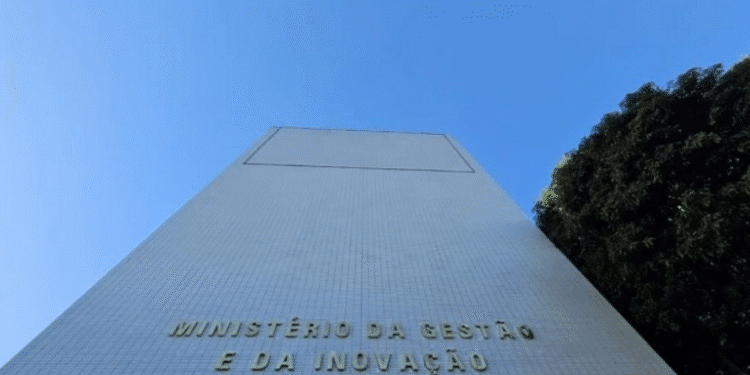 Foto da fachada de um edifício alto com a inscrição "MINISTÉRIO DA GESTÃO E DA INOVAÇÃO EM SERVIÇOS PÚBLICOS" contra um céu azul claro e árvores ao lado.
