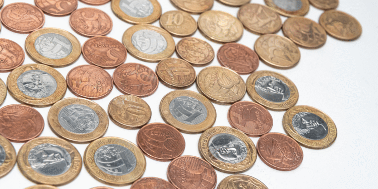 Diversas moedas brasileiras de diferentes valores e cores espalhadas sobre superfície branca
