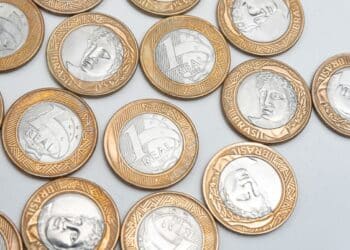 Conjunto de moedas brasileiras de 1 Real espalhadas sobre uma superfície branca, representando colecionismo, valor e numismática.