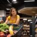 Mulher sorrindo em um corredor de supermercado com o carrinho cheio de frutas e verduras, celebrando a notícia de que os preços caíram com a desaceleração da inflação.