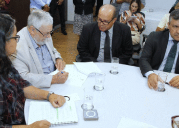 Pessoas em reunião sentadas à mesa com documentos, copos de água e assinatura sendo feita