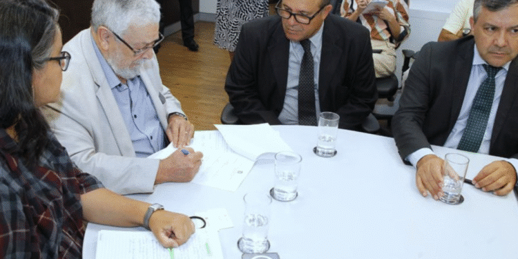 Pessoas em reunião sentadas à mesa com documentos, copos de água e assinatura sendo feita