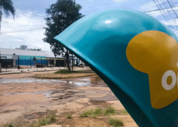 Cabine telefônica azul da Oi em rua com chão de terra batida próxima a prédio público.