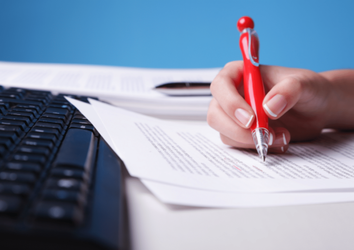 Foto close-up de uma mão feminina segurando uma caneta vermelha e escrevendo em um documento de papel sobre uma mesa, com teclado de computador ao lado e fundo azul.