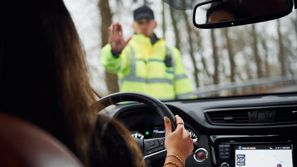 Evite infrações não óbvias e multas inesperadas Um agente de trânsito em colete refletivo sinaliza para uma motorista parar seu veículo, simbolizando a fiscalização e a necessidade de atenção às infrações do CTB.