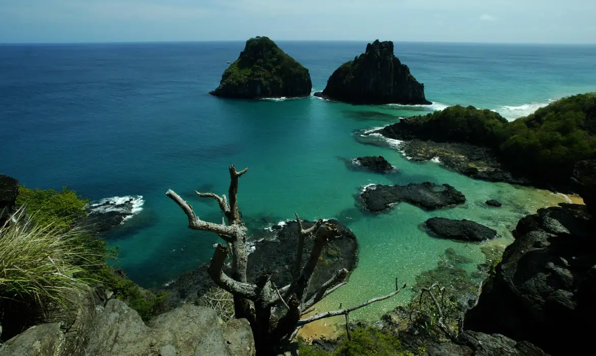 Vista aérea da baía com águas cristalinas, duas ilhas rochosas e vegetação em Fernando de Noronha
