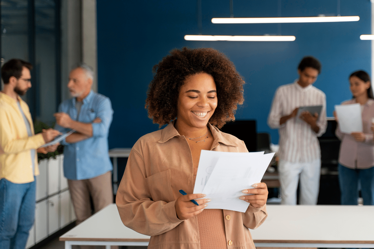 Mulher sorridente lendo papéis em mãos com colegas conversando ao fundo em ambiente de trabalho.