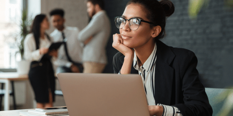 Mulher jovem usando óculos, sentada em mesa com laptop, olhando pensativa, com colegas ao fundo no escritório