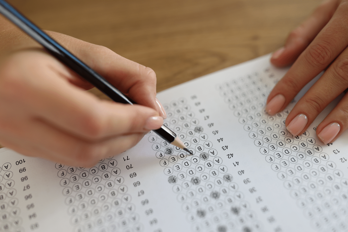 Concurso Unificado PE Mãos com unhas feitas preenchendo uma prova de múltipla escolha com lápis preto em folha branca