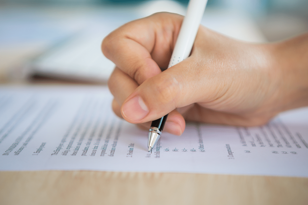 Concursos Prefeituras SP  Mão segurando caneta branca preenchendo formulário impresso em mesa de madeira