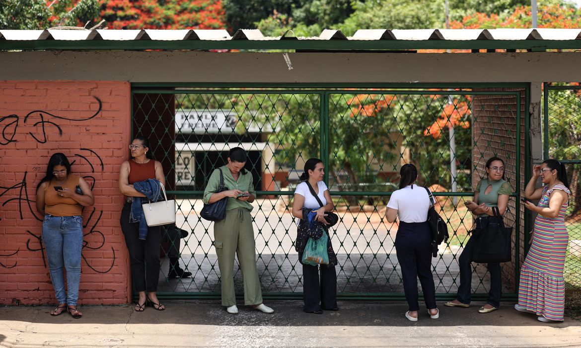 Candidatas utilizando celulares enquanto aguardam em frente ao portão de entrada de um local para fazera a Prova Nacional Docente (PND).