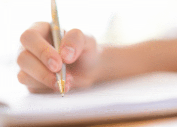 Mão segurando caneta dourada e escrevendo em folhas de papel branco sobre superfície clara