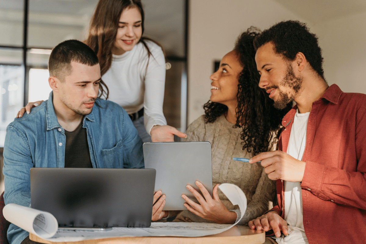 Quatro jovens trabalhando juntos em mesa, usando laptop e tablet para colaboração.