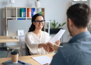 Mulher sorridente apertando a mão de um entrevistador, simbolizando o sucesso em uma entrevista de emprego após um currículo atrativo.