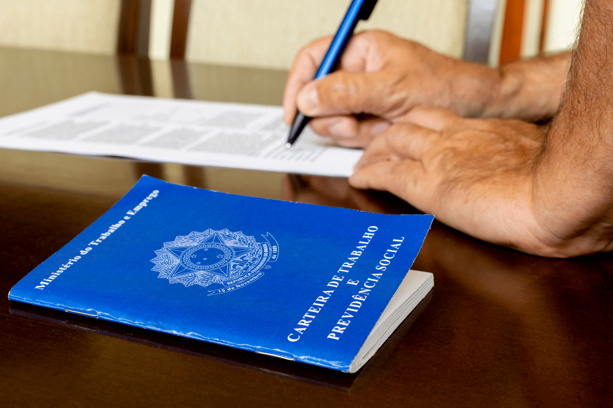 Vagas de emprego Mãos masculinas assinando documento ao lado de carteira de trabalho azul sobre mesa de madeira