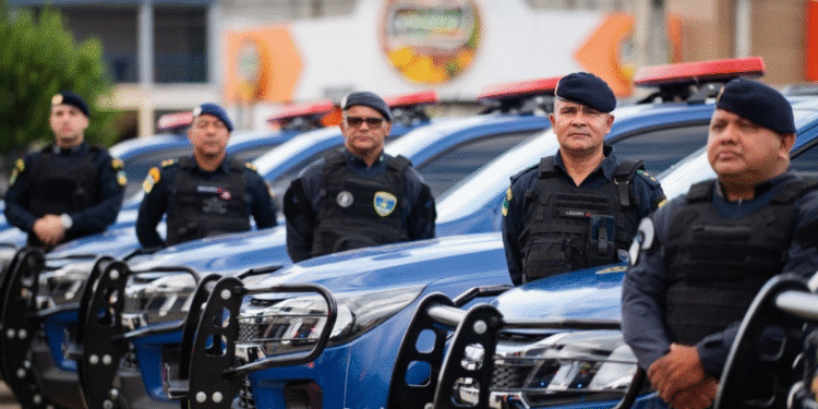 Grupo de guardas municipais em pé ao lado de viaturas policiais azuis alinhadas em fila, todos com uniformes e coletes táticos, usando boinas.