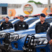Grupo de guardas municipais em pé ao lado de viaturas policiais azuis alinhadas em fila, todos com uniformes e coletes táticos, usando boinas.