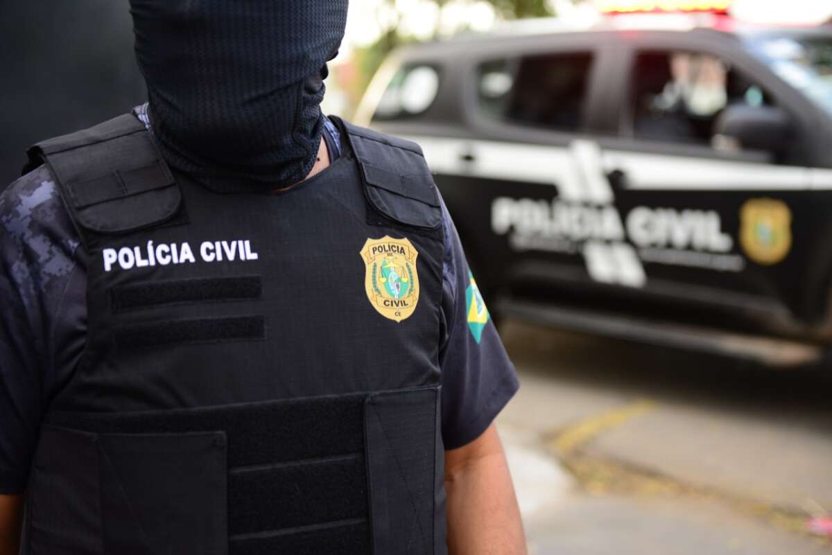 Agente da Polícia Civil usando colete balístico preto e máscara, com viatura ao fundo