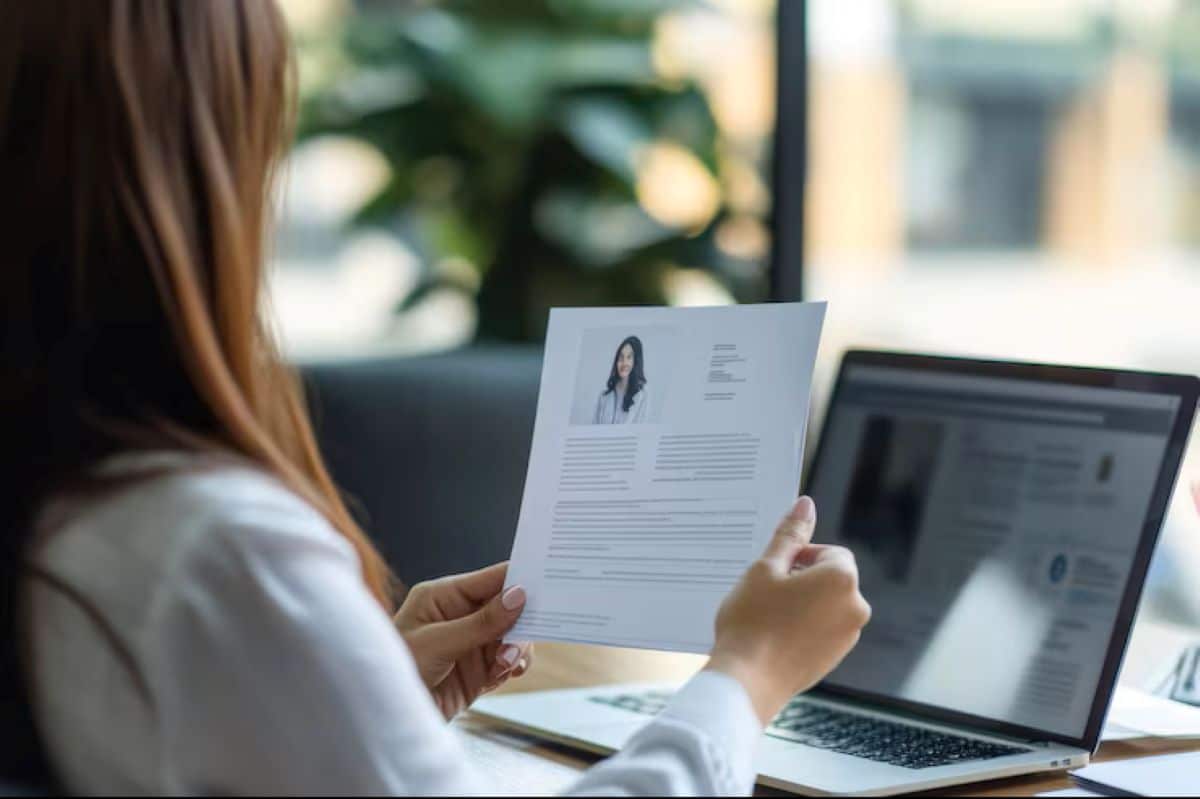 Pessoa revisando um currículo impresso com atenção, enquanto um notebook está aberto ao lado, representando a importância da revisão antes de se candidatar.