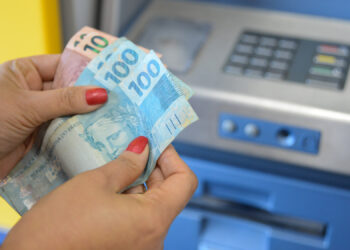 Mãos femininas segurando cédulas de reais próximo a um caixa eletrônico azul