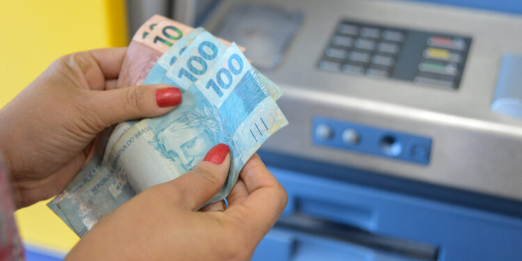Mãos femininas segurando cédulas de reais próximo a um caixa eletrônico azul