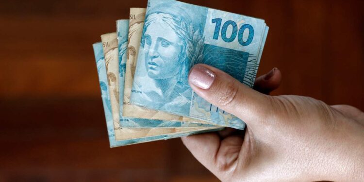 Mão feminina segurando notas de dinheiro em reais, incluindo uma nota de 100 reais e várias notas de 50 reais