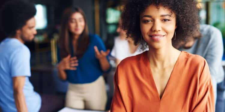 Profissional confiante em ambiente de trabalho moderno com colegas interagindo ao fundo.