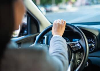 Mulher ao volante em aula de direção com instrutor avaliando e documento da CNH ao fundo.