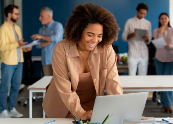 Jovem profissional sorridente trabalhando em notebook em escritório moderno com equipe colaborando ao fundo.