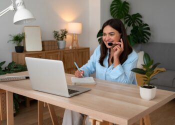 Profissional sorridente realizando atendimento remoto com headset em escritório doméstico aconchegante fazendo anotações durante videochamada.