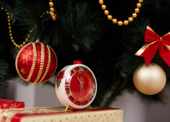 Relógio decorativo de Natal ao lado de bolas natalinas vermelhas e douradas em árvore de Natal.