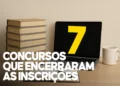 Notebook aberto exibindo o número 7 amarelo na tela, posicionado ao lado de uma pilha de livros beges, ilustrando o encerramento de 7 concursos de prefeituras.