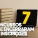 Notebook aberto exibindo o número 7 amarelo na tela, posicionado ao lado de uma pilha de livros beges, ilustrando o encerramento de 7 concursos de prefeituras.