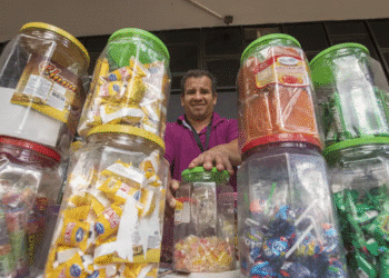 Homem sorridente atrás de potes transparentes cheios de doces embalados à venda em mercado informal.