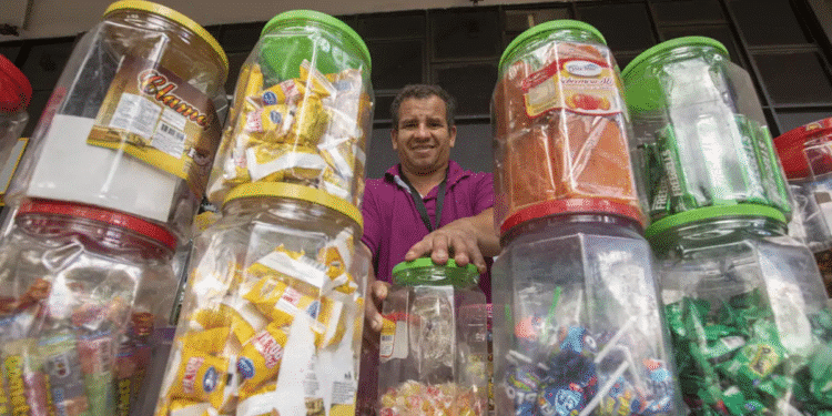Homem sorridente atrás de potes transparentes cheios de doces embalados à venda em mercado informal.