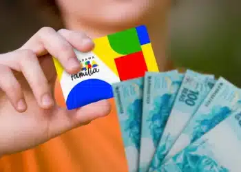 Mão segurando cartão colorido do Programa Auxílio Família e notas de 100 reais