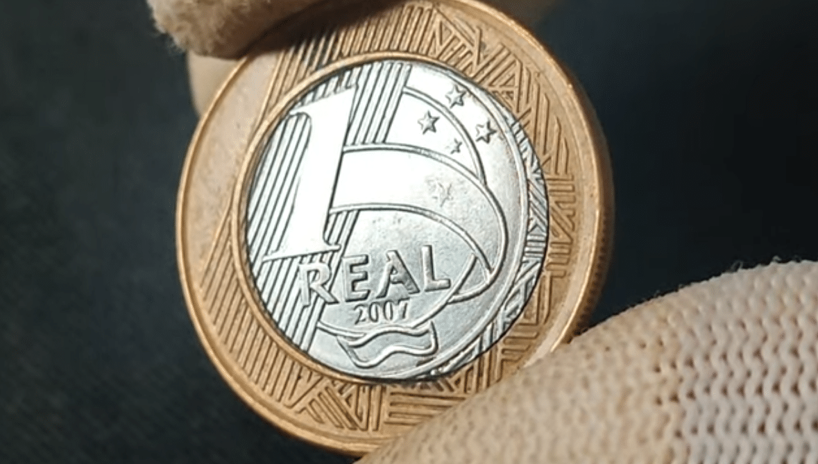 Moeda de R$ 1 do ano de 2007 sendo examinada de perto, ilustrando um possível defeito de núcleo deslocado, que eleva seu valor para colecionadores até R$ 260, dependendo da conservação.