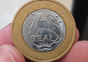 Close-up de uma mão segurando a face de valor de uma moeda de 1 real do ano de 2004, com o núcleo de aço inox e anel de bronze.