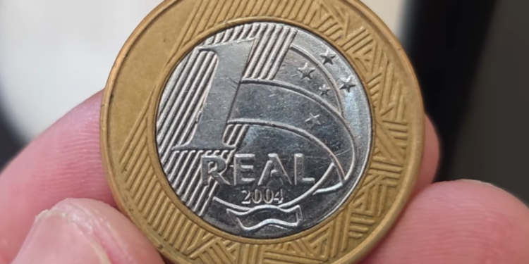 Close-up de uma mão segurando a face de valor de uma moeda de 1 real do ano de 2004, com o núcleo de aço inox e anel de bronze.