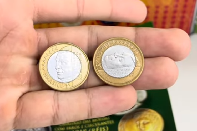Mão segurando duas moedas de 1 Real de Juscelino Kubitschek lado a lado, mostrando a diferença de alinhamento do reverso entre uma peça comum e uma com erro.