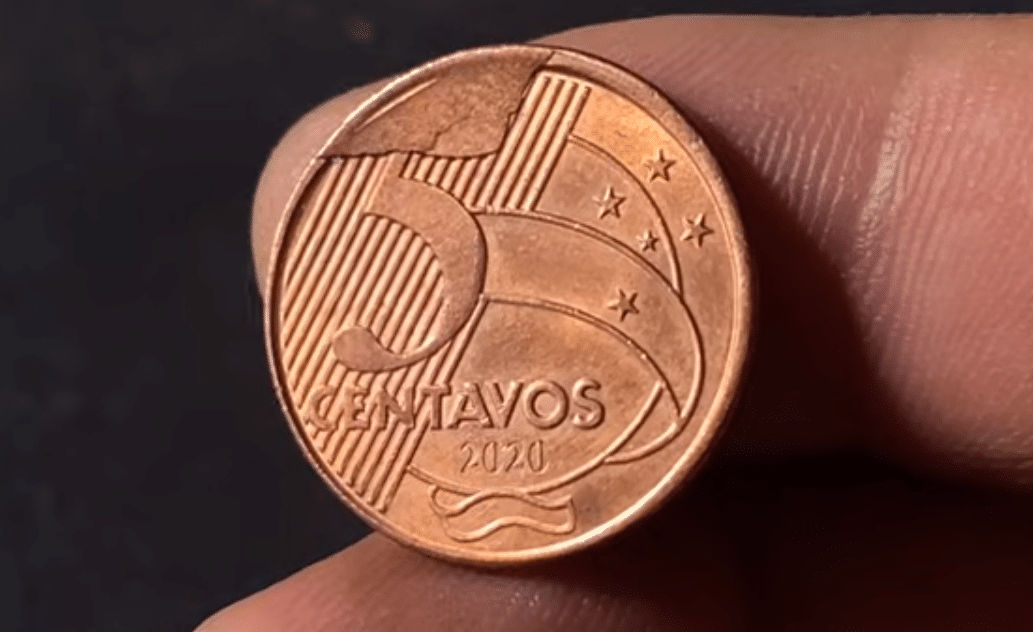 moeda 5 centavos 2020 detalhe erro cunho quebrado Close de uma moeda de 5 centavos de 2020 segurada por dedos, apresentando uma falha de metal em relevo na borda superior, característica do erro de cunho quebrado.