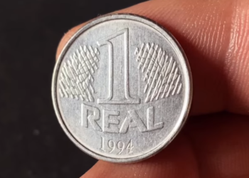 Close nítido de uma moeda de 1 Real de aço inoxidável do ano 1994, segurada por dedos, destacando o valor de face e a data na parte inferior.
