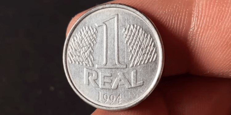 Close nítido de uma moeda de 1 Real de aço inoxidável do ano 1994, segurada por dedos, destacando o valor de face e a data na parte inferior.