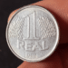 Close nítido de uma moeda de 1 Real de aço inoxidável do ano 1994, segurada por dedos, destacando o valor de face e a data na parte inferior.