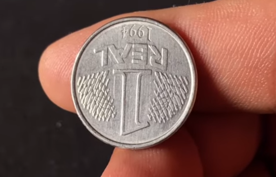 Close de uma moeda de 1 Real de 1994 sendo segurada entre os dedos, exibindo o valor de face e o ano de ponta-cabeça, demonstrando o erro de reverso invertido.