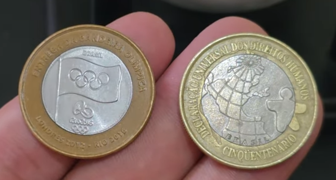 Close-up de duas moedas de 1 real lado a lado: à esquerda a moeda DH de 1998 e à direita a moeda da Bandeira Olímpica de 2012, ambas em estado de conservação Flor de Cunho.